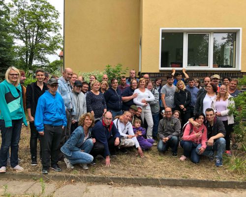 IMG_9279 Eine große Gruppe Menschen vor einem Gebäude mit gelber Fassade. Alle lachen und schauen in die Kamera
